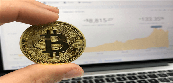 Bitcoin zwischen Fingern vor einem Laptop-Screen mit Kursen